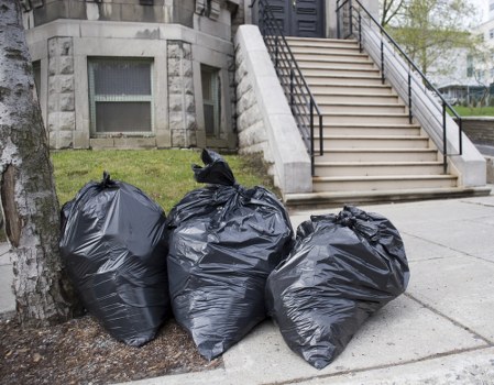 Three large black plastic rubbish bags, filled and tightly tied at the top, are positioned on the pavement next to a tree trunk in front of a residential building in Regents Park. The bags appear to contain outdoor waste, possibly household or garden refuse, and are placed in close proximity to each other, partially leaning against the tree and on the concrete surface. In the background, there is a set of concrete stairs with black metal handrails leading up to an entrance of the building, which has a grey stone façade and small windows with external security grilles. The scene is set on a typical urban street in central London, near the postcode area of NW1, with natural light suggesting a daytime setting. This image reflects a typical outdoor rubbish disposal scene, relevant to commercial waste removal services provided by Commercial Waste Regents Park, serving properties in the local area with residential or commercial waste clearance needs.