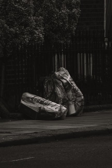 A large outdoor pile of used books stacked haphazardly on the pavement near a weathered wooden fence and a decorative street lamp post. The books vary in size, color, and condition, with some stacked 