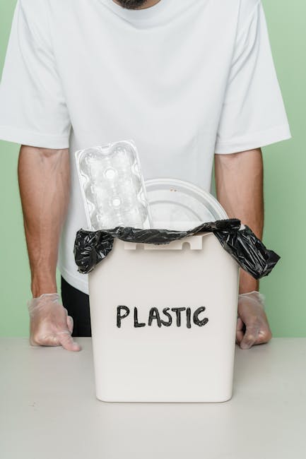 A group of large black plastic garbage bags filled with waste materials are placed on the pavement beside a black metal fence with vertical bars. The bags appear to contain household or garden rubbish